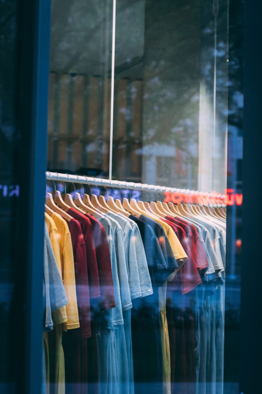 A rail of coloured t-shirts on wooden hangers seen through a shop window.